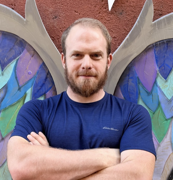Gideon Kai, arms crossed, in front of a painted mural of wings